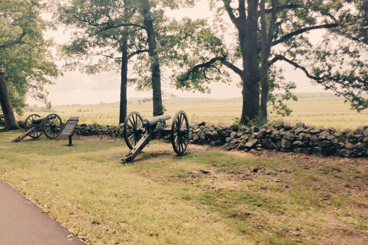 Historic battlefield landscape featuring vintage cannons lined up along a stone wall surrounded by lush trees and greenery, depicting a serene and preserved Civil War site.