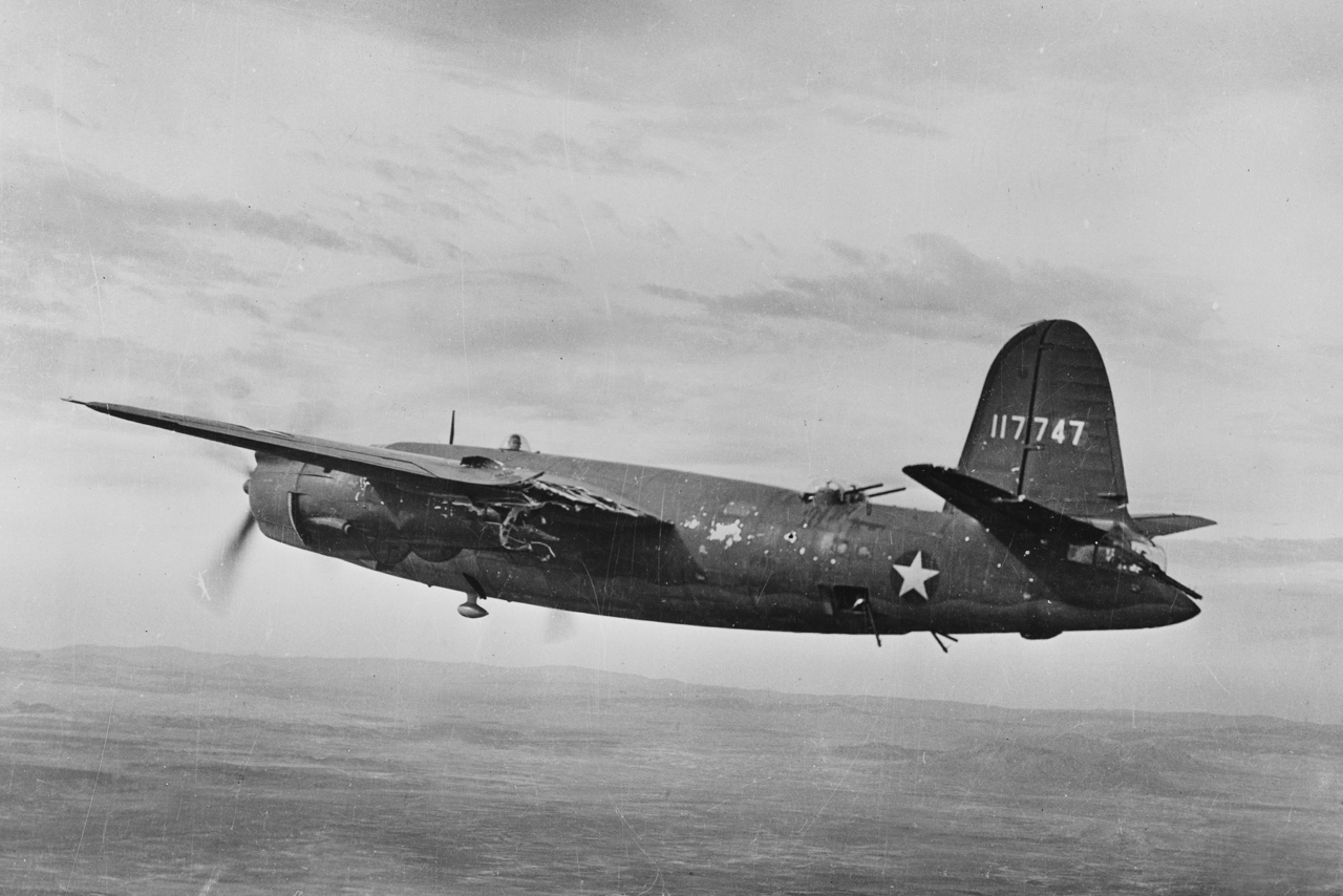 World War II American bomber aircraft with battle damage to its left wing flying over a landscape, displaying US Air Force star insignia and tail number 117747.