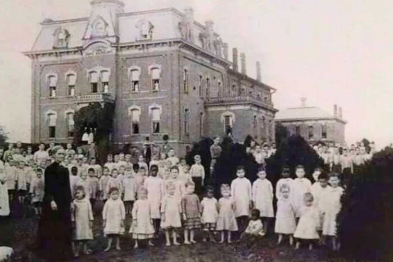 Historic black and white photo showing a large group of children and adults posing outside a brick orphanage or boarding school building from the late 19th or early 20th century.