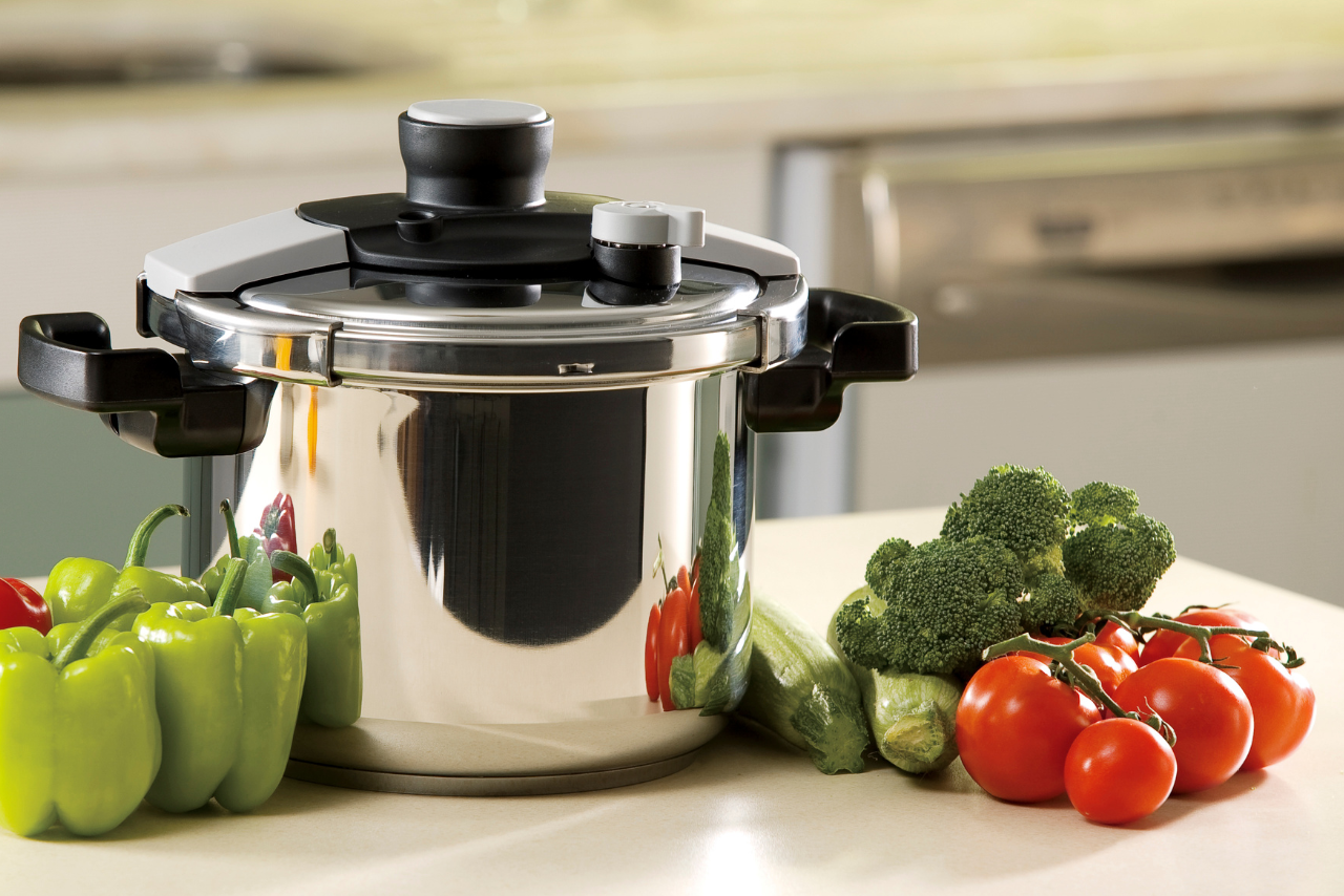 A stainless steel pressure cooker on a kitchen counter with green bell peppers, zucchini, broccoli, and tomatoes beside it.