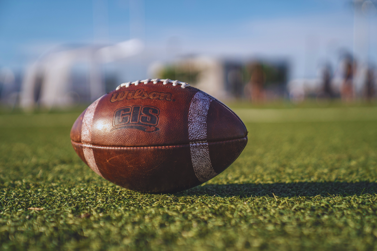 A close-up view of a brown leather American football resting on a grassy field.   The football is slightly angled, with the stitching and the "GIS" logo clearly visible.   The background is out of focus, showing a portion of a sports field and a partly cloudy sky.   The grass is a vibrant green, and the lighting suggests a sunny day. A shadow is cast by the football onto the grass.