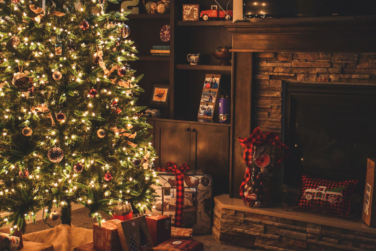 A warmly lit Christmas tree, adorned with ornaments and lights, stands beside a cozy fireplace. Gifts wrapped in festive paper are placed underneath.