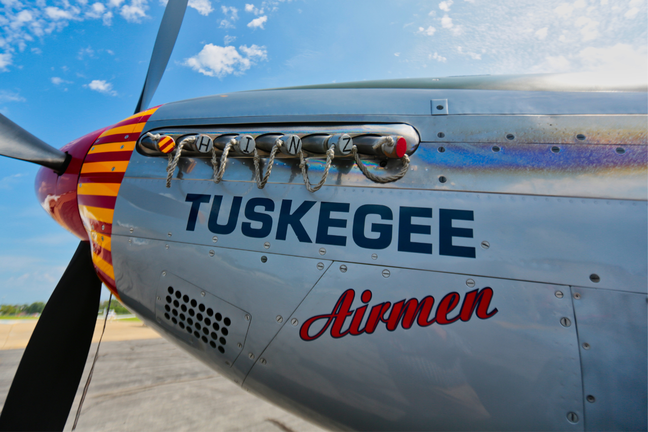 Close-up of a vintage airplane's nose with "Tuskegee Airmen" painted on the side. The propeller and engine detail convey historical significance. Bright sky in background.