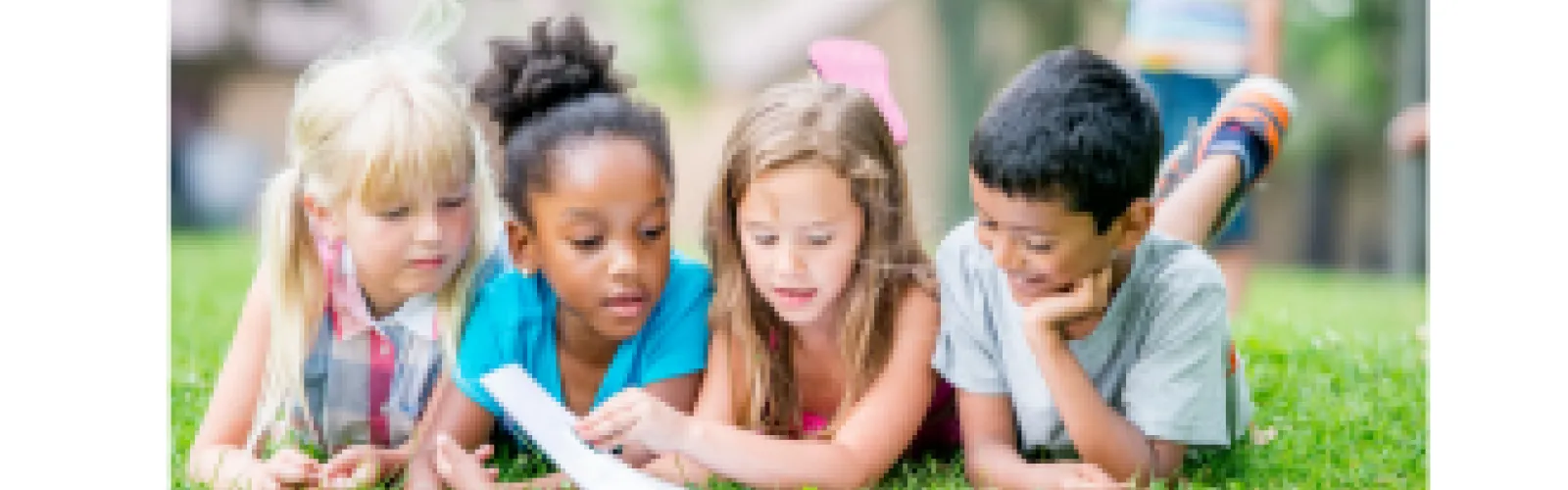 Children reading a book together while lying on grass in a park, diverse group of kids enjoying outdoor learning and education