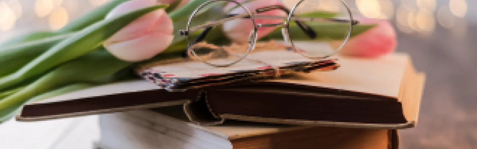 Vintage eyeglasses resting on stacked books with pink tulips and soft bokeh lights in the background, creating a cozy reading scene.